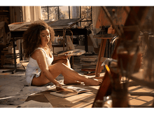 Charger l&#39;image dans la galerie, Femme dans un atelier d&#39;art, tenant une palette, entourée de fournitures artistiques. Lumière solaire.
