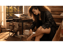 Charger l&#39;image dans la galerie, Une femme en robe et veste noires, qui applique de la lotion sur sa jambe, dans un studio.
