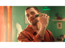 Charger l'image dans la galerie, Un homme se rase le visage avec une tondeuse électrique. Il porte une chemise orange. Fond vert.
