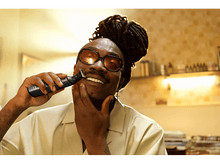 Charger l&#39;image dans la galerie, Un homme noir se rase la moustache avec une tondeuse noire. Il sourit et porte des lunettes.

