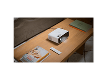 Charger l'image dans la galerie, Projecteur blanc sur table en bois avec livres et télécommande. Canapé en arrière-plan.
