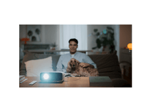 Charger l'image dans la galerie, Un homme avec un chien sur un canapé, projecteur sur une table. Le projecteur brille. Livres et télécommande sur la table.
