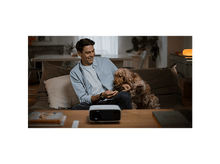 Charger l'image dans la galerie, Un homme sourit, caressant un chien brun sur un canapé. Un projecteur est sur la table. Livres et décor au fond.
