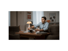 Charger l'image dans la galerie, Un homme ajuste un projecteur sur une table. Livres et canapé au premier plan. Art et lampe en arrière-plan.
