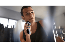 Charger l&#39;image dans la galerie, Homme se rasant avec un rasoir électrique noir et bleu. Il regarde en l&#39;air. L&#39;arrière-plan est flou.
