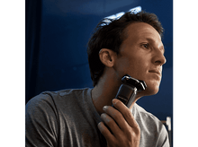 Charger l&#39;image dans la galerie, Un homme se rase le visage avec un rasoir électrique. Il porte une chemise grise.
