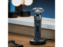 Charger l&#39;image dans la galerie, Un rasoir électrique noir et bleu sur une base de chargement. Fond de table en bois.
