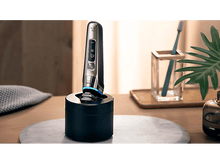 Charger l&#39;image dans la galerie, Rasoir électrique dans socle de charge, à côté d&#39;une brosse à dents, sur marbre.
