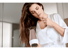 Charger l&#39;image dans la galerie, Femme séchant ses cheveux avec un sèche-cheveux or rose dans une salle de bain, portant une robe blanche.
