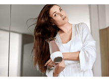Charger l&#39;image dans la galerie, Une femme en peignoir blanc sèche ses longs cheveux bruns dans une salle de bain.
