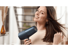 Charger l&#39;image dans la galerie, Femme sourit utilisant un sèche-cheveux bleu foncé, cheveux au vent. Escalier et plantes en arrière-plan.
