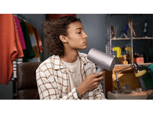 Charger l&#39;image dans la galerie, Un jeune homme utilise un sèche-cheveux. Il est dans une pièce avec des vêtements et une lampe en arrière-plan.
