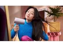 Charger l&#39;image dans la galerie, Femme sèche ses cheveux au sèche-cheveux. Chemise bleue, pantalon rose, miroir et plante.
