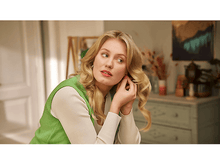 Charger l&#39;image dans la galerie, Femme blonde ajustant une boucle d&#39;oreille, arrière-plan flou d&#39;une pièce avec des œuvres d&#39;art et des arrangements floraux.
