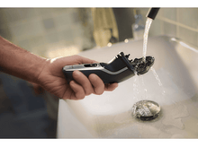 Charger l&#39;image dans la galerie, Une main tenant un rasoir électrique noir et argenté sous l&#39;eau courante dans un lavabo blanc.
