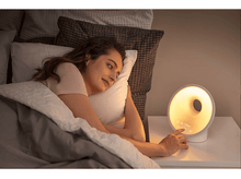 Charger l&#39;image dans la galerie, Une femme est allongée dans son lit, tendant la main vers un réveil blanc. La lumière brille de l&#39;horloge vers la table de chevet.
