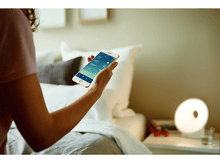Charger l&#39;image dans la galerie, Personne tenant un smartphone affichant une appli domotique, lampe sur table de chevet.
