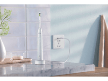 Charger l&#39;image dans la galerie, Une brosse à dents électrique sur une base de chargement, près d&#39;un mur avec une prise et une serviette.
