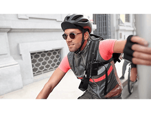 Charger l&#39;image dans la galerie, Un homme à vélo portant un casque noir, des lunettes de soleil et un gilet avec une radio. Le fond est une ville.
