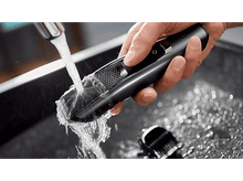 Charger l&#39;image dans la galerie, Une main rince un rasoir électrique noir et gris sous l&#39;eau courante dans un évier.
