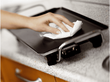 Charger l&#39;image dans la galerie, Une main essuie une plaque de cuisson noire avec une serviette en papier blanche.
