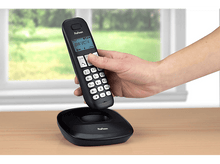 Charger l&#39;image dans la galerie, Un téléphone sans fil noir sur une base de chargement, tenu par une main, devant une fenêtre.
