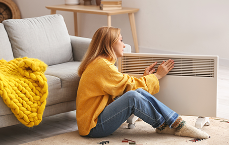 Une jeune femme se réchauffe auprès d'un radiateur mobile électrique dans son salon.
