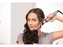 Charger l&#39;image dans la galerie, Femme se bouclant les cheveux avec un fer à friser, portant un gant, regardant la caméra.
