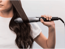 Charger l&#39;image dans la galerie, Une femme se lisse les cheveux foncés avec un fer à lisser gris.
