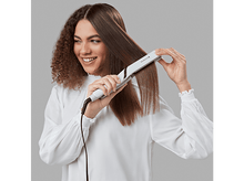 Charger l&#39;image dans la galerie, Femme aux cheveux longs se lisse les cheveux avec un fer plat, souriant au spectateur.
