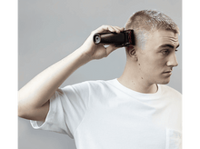 Charger l&#39;image dans la galerie, Un homme se coupe les cheveux avec une tondeuse noire. Il la tient contre sa tête.
