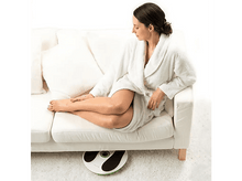 Charger l'image dans la galerie, Une femme en peignoir blanc assise sur un canapé blanc, avec un masseur de pieds au sol.

