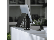 Charger l&#39;image dans la galerie, Une caméra Ring noire est posée sur un bureau blanc. Un ordinateur portable et un téléphone sont en arrière-plan.
