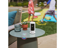 Charger l&#39;image dans la galerie, Une caméra Ring est posée sur une table avec un verre, une plante et des enfants jouant en arrière-plan avec des bulles.
