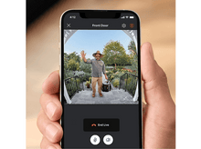 Charger l&#39;image dans la galerie, Une personne tenant un téléphone affichant un appel vidéo depuis une porte d&#39;entrée. Un homme fait signe.
