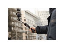 Charger l&#39;image dans la galerie, Personne tenant une perche à selfie avec un téléphone, prenant une photo. Bâtiments en arrière-plan.
