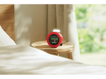 Charger l&#39;image dans la galerie, Réveil rouge sur une table de chevet. Indique l&#39;heure, 12h00. Lit et fenêtre en arrière-plan.
