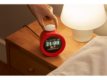 Charger l&#39;image dans la galerie, Une main ajuste un réveil rouge sur une table de chevet avec une lampe. L&#39;horloge affiche l&#39;heure.
