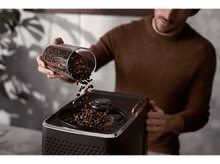 Charger l&#39;image dans la galerie, Un homme versant des grains de café dans une machine à café noire.
