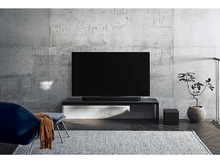 Charger l&#39;image dans la galerie, Salon avec TV sur meuble, barre de son, fauteuil bleu, tapis et plante.
