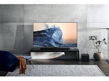 Charger l&#39;image dans la galerie, Une grande télévision affiche une vague. Une barre de son est en dessous. Une chaise bleue et un tapis sont visibles.
