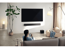 Charger l&#39;image dans la galerie, Une personne assise sur un canapé, regardant une télévision. Une barre de son, un caisson de basses et une plante sont visibles.
