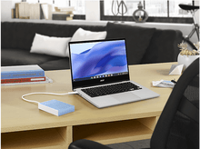 Charger l&#39;image dans la galerie, Ordinateur portable ouvert sur un bureau en bois avec un disque dur externe bleu. Livres et une plante en arrière-plan.
