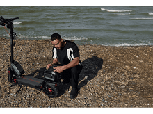 Charger l&#39;image dans la galerie, Homme agenouillé près d&#39;un scooter noir sur une plage rocheuse. Océan en arrière-plan, eau verte.
