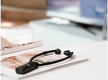 Charger l&#39;image dans la galerie, Aide auditive noire à tour de cou sur un magazine. Table blanche et arrière-plan flou.
