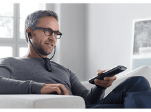 Charger l&#39;image dans la galerie, Un homme avec des lunettes tient une télécommande, portant un appareil autour du cou, assis sur un canapé blanc.
