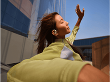 Charger l&#39;image dans la galerie, Femme utilisant des écouteurs à l&#39;extérieur. Lève la main et sourit. Devant un bâtiment ensoleillé.
