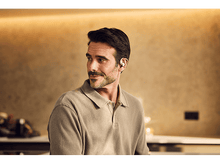 Charger l&#39;image dans la galerie, Homme portant un écouteur sans fil, regardant ailleurs. Arrière-plan flou d&#39;une cuisine ou salle à manger.
