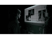 Charger l&#39;image dans la galerie, Homme regardant des radiographies sur un mur. Un squelette à droite. Pièce sombre.
