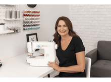 Charger l&#39;image dans la galerie, Une femme souriante à côté d&#39;une machine à coudre blanche. Elle est dans une pièce bien éclairée avec des bobines de fil.
